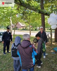Mehrere Besucherinnen und Besucher stehen auf einer Grünfläche unter schattigen Bäumen am Malteser-Stand und nehmen an einem Spielangebot teil. Rechts im Bild sind weiße Malteser-Luftballons zu sehen, im Hintergrund eine Straße sowie Fahrräder.
