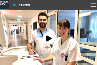 Zwei Pflegekräfte an einem Stationsarbeitsplatz mit Dokumenten und Computern in einem hellen Flur.