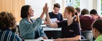 Eine Frau und ein Mädchen schlagen gemeinsam ein. Sie sitzen an einem Tisch im Klassenraum.