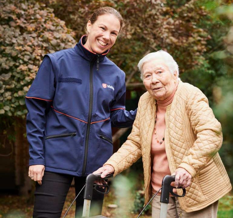 Eine junge Frau in Malteser Jacke und eine ältere Dame mit Rollator stehen im Grünen und lächeln.
