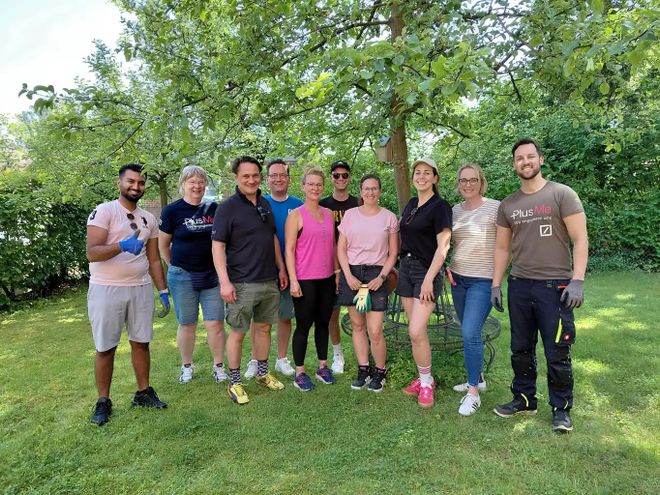 Gruppenfoto im Garten des Malteser Hospiz-Zentrums: Das Team der Deutschen Bank Filiale Volksdorf und Mitarbeitende des Malteser Hospiz-Zentrums stehen gemeinsam auf einer Rasenfläche unter einem großen Baum. Im Vordergrund liegt ein Hund auf dem Gras.