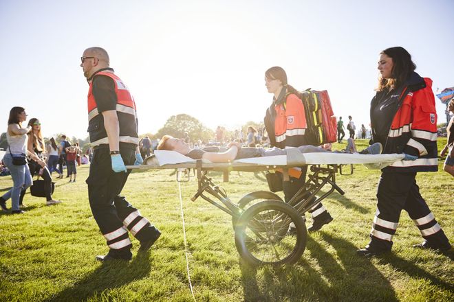 Mehrere Sanitätsdienstkräfte transportieren eine Person auf einer Trage über ein Veranstaltungsgelände im Freien. Im Hintergrund sind Besucherinnen und Besucher auf einer großen Wiese bei sonnigem Wetter zu sehen.