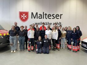 Auf dem Foto sind die Teilnehmenden und Dozenten des Lehrgangs in der Fahrzeughalle zu sehen. Im Hintergrund an der Wand kann man das Logo der Malteser erkennen.