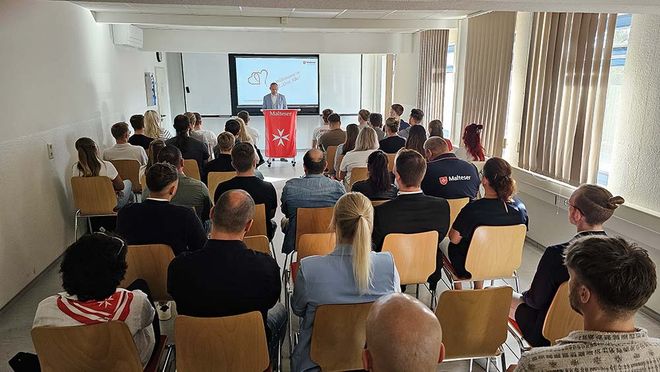 Viele Menschen sitzen in einem Raum. Ein Mann hält eine Rede.