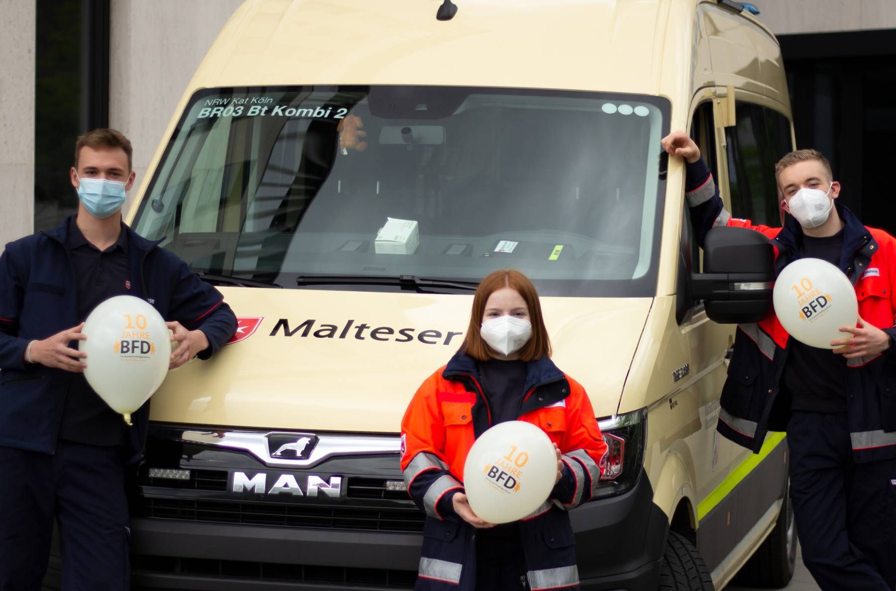 Eine junge Frau und zwei junge Männer halten Luftballons vor einem Kleinbus in die Höhe