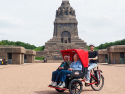 Eine Seniorin und ein Senior sitzen in der Rikscha. Ein Malteser Mitarbeitende sitzt auf dem Sattel. Im Hintergrund ist das Völkerschlachtdenkmal zu sehen.