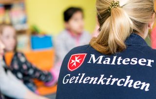 Das Bild zeigt eine Szene in einem Schulraum der Offenen Ganztagsbetreuung. Eine Frau steht mit dem Rücken zur Kamera und trägt ein dunkelblaues Shirt mit der Aufschrift 'Malteser Geilenkirchen'. Im Hintergrund sind Kinder zu sehen, die unscharf und beim Spielen sind. Der Raum ist mit Tischen und Stühlen eingerichtet, und an den Wänden hängen Poster und Kunstwerke von Kindern. Es scheint eine heitere und entspannte Atmosphäre im Raum zu herrschen