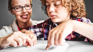 Frau sitzt hinter einem Jungen, der in Blindenschrift liest.