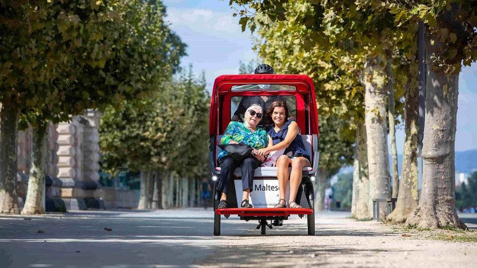 Eine ältere Frau und ein junges Mädchen sitzen in einer Rikscha, die sie durch eine Allee fährt.