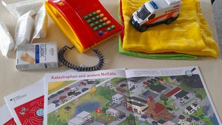 Kinderbuch, das eine Rettungsstelle von oben zeigt, Verbandszeug, ein buntes Spielzeugtelefon und ein Modell eines Rettungswagens.