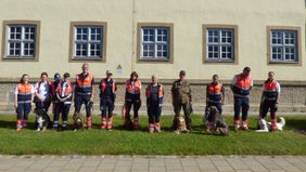 12 Männer und Frauen und einige Hunde stehen aufgereiht vor einem Kasernengebäude.