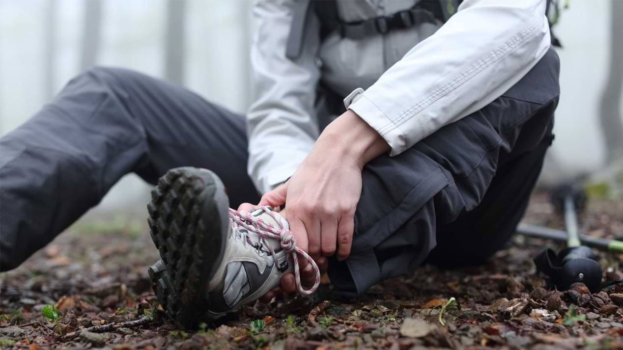 Ein Mann in Outdoor-Kleidung sitzt auf einem Waldboden und hält sich den Fußknöchel.
