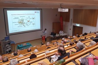 Zuschauer verfolgen in einem Hörsaal eine medizinische Präsentation mit Vortragenden und Projektionsfläche vorne.