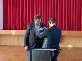 Ein Mann steckt einem anderen eine Ehrennadel an. 