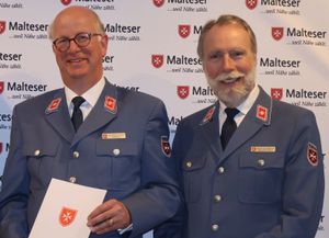 Zwei Männer in grauer Malteser Uniformjacke stehen vor einer weißen Wand mit vielen Malteser Logos