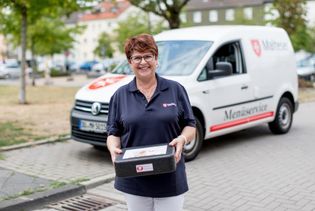 Mitarbeiterin des Malteser Menueservice trägt eine Warmhaltebox und lächelt in die Kamera.