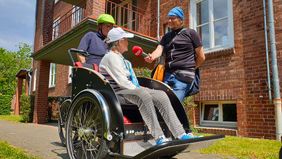 Ein Mann hält ein Mikrofon. In dieses Mikrofon spricht eine Frau, die in einer Fahrradrikscha sitzt. 