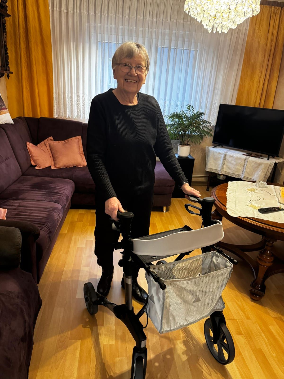 eine ältere Dame mit Rollator steht in der Mitte ihres Wohnzimmers und lächelt in die Kamera