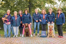 Die Helfenden des Malteser Besuchs- und Begleitungsdienstes stehen gemeinsam mit ihren Hunden vor einem Baum. 