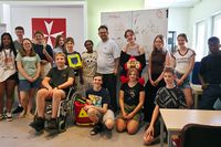 Gruppe von etwa 14 Jugendlichen und einer erwachsenen Person in einem Klassen- oder Schulungsraum. Die Gruppe steht und sitzt für ein gemeinsames Foto vor einem Whiteboard. Im Vordergrund sind mehrere Gegenstände zur Erste-Hilfe-Ausbildung zu sehen, darunter ein Trainings-Defibrillator, ein Rettungsrucksack und eine Puppe zur Übung von Rettungsmaßnahmen. Eine Person sitzt in einem Rollstuhl. An der Wand hängt ein rotes Banner mit dem weißen Malteser-Kreuz. Helle Tische und Stühle stehen im Raum. Das Foto zeigt eine Schulungs- oder Projektgruppe im Bereich Erste Hilfe und Rettungsdienst.