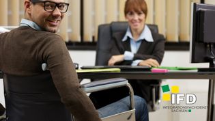 Mann im Rollstuhl mit braunem Pullover und Brille sitzt einem Geschäftsfrau gegenüber, die an einem Schreibtisch sitzt.