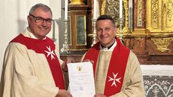 Domkapitular Hans-Georg Platschek und Prodekan Fabian Orsetti, beide mit liturgischem Gewand und Stola mit Malteserkreuz, halten gemeinsam eine Urkunde. 