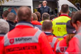 Ein Mann spricht am Mikrofon. Zahlreiche Menschen in Einsatzkleidung stehen vor ihm.