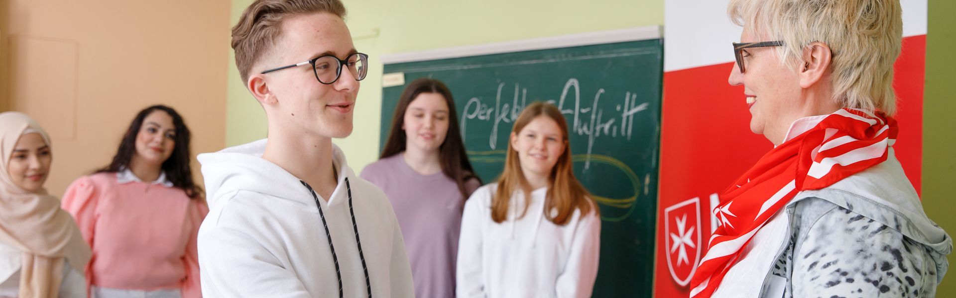 Ein Schüler schüttelt die Hand einer Benimmtrainerin in einem Klassenraum.
