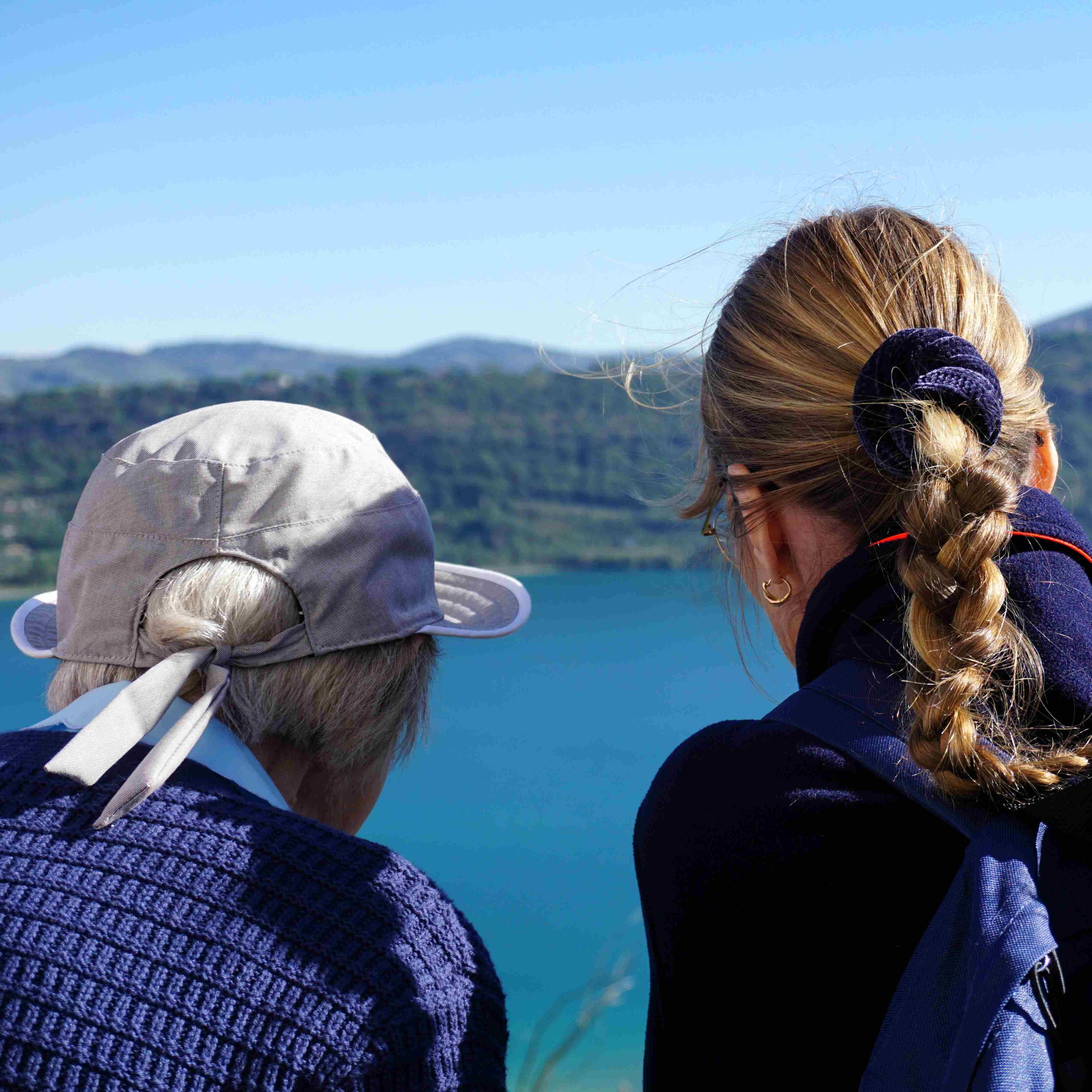 Zwei Personen von hinten, eine mit hellgrauem Hut, die andere mit geflochtenem Haar, blicken auf einen See und bewaldete Hügel in Castel Gandolfo.