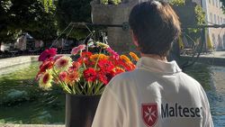 Eine Frau mit Malteser-Shirt (von hinten) und Blumen steht an einem Brunnen in Bad Reichenhall.