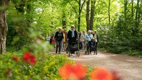 Eine Gruppe von älteren Leuten mit Rollatoren oder andern Gehilfen geht mit Ehrenamtlichen vom Malteser Hilfsdienst durch den grünen Britzer Garten.