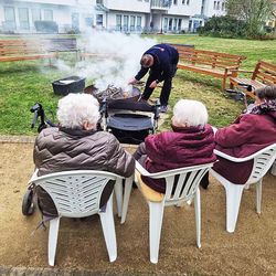 Seniorinnen sitzen an einer Feuerschale. In dieser zündet ein Mann gerade ein Feuer an.