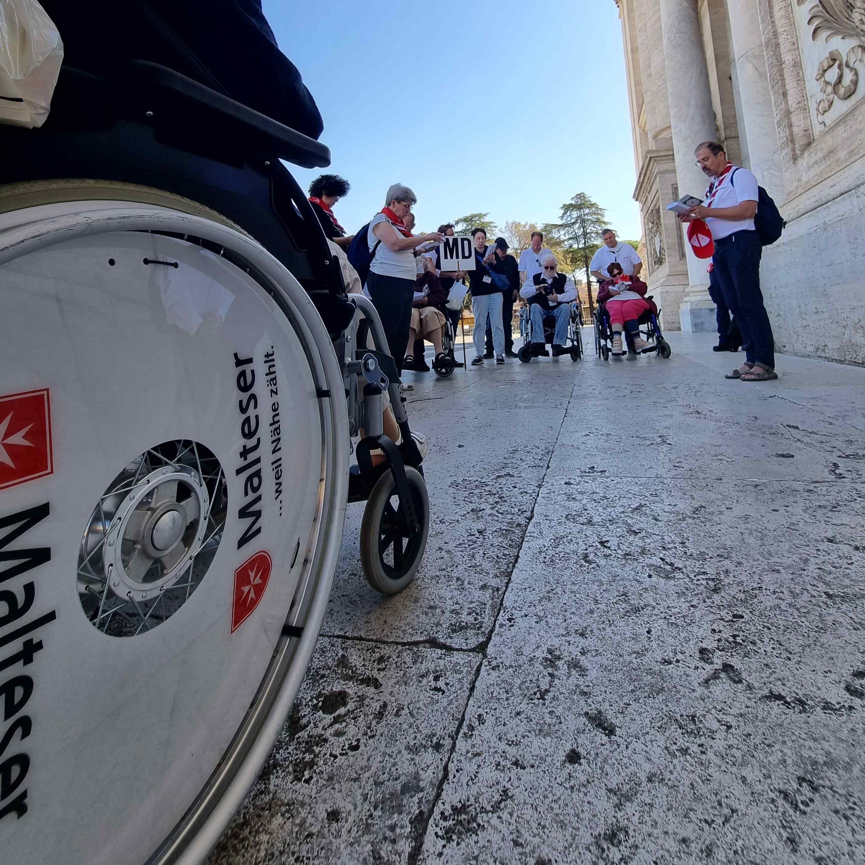 Rollstuhl mit Aufschrift 'Malteser' auf dem Rad, im Hintergrund Menschen vor einer historischen Fassade bei der Romwallfahrt.