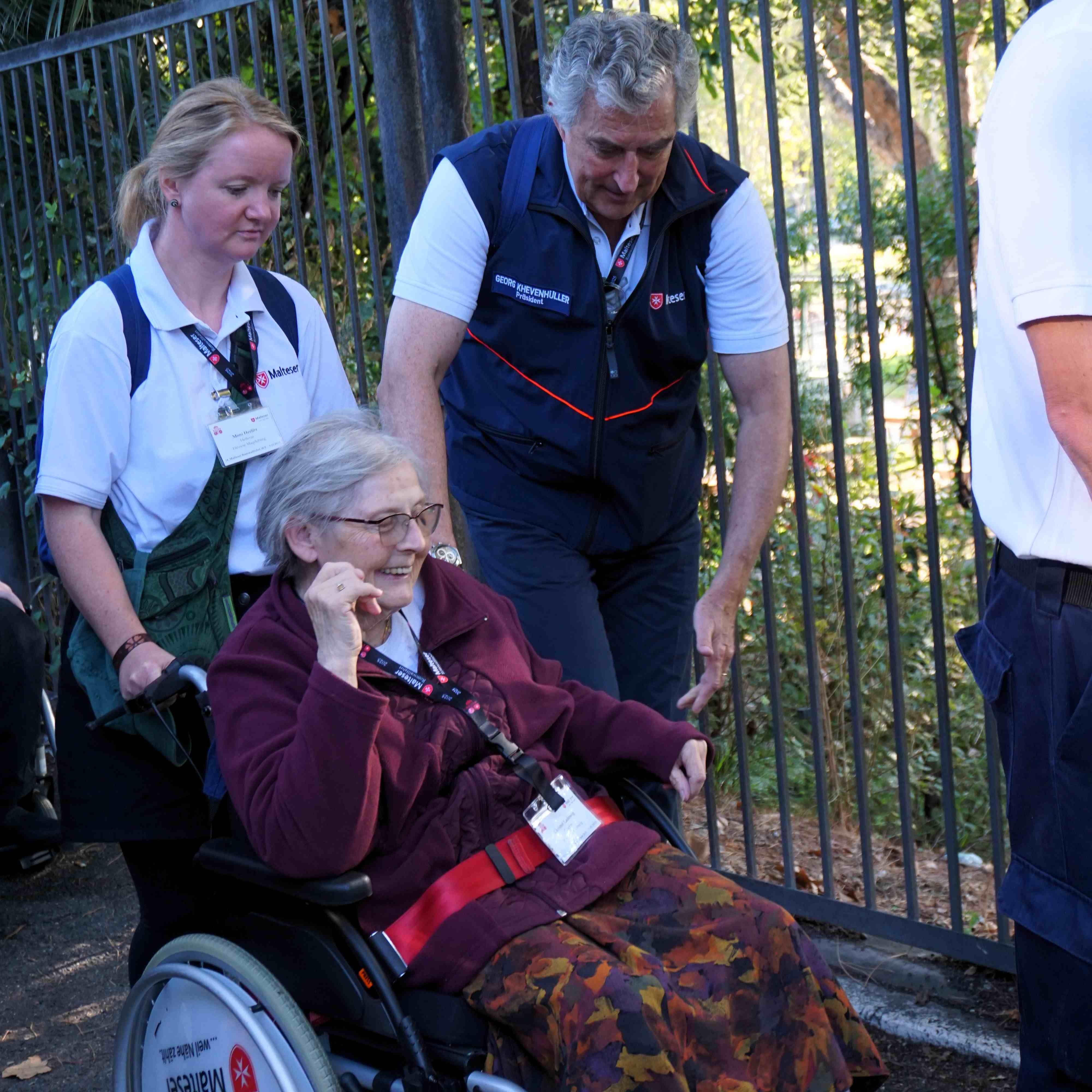 Person im Rollstuhl mit rotem Pullover und gemusterter Hose wird von zwei Personen begleitet, eine trägt dunkelblaue Weste, im Hintergrund Metallzaun.