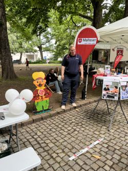 Ein Stand ist aufgebaut auf der Straße mit Informationsmaterila und Spiele für die Kinder. Im Hintergrund steht eine Malteser Mitarbeiterin.