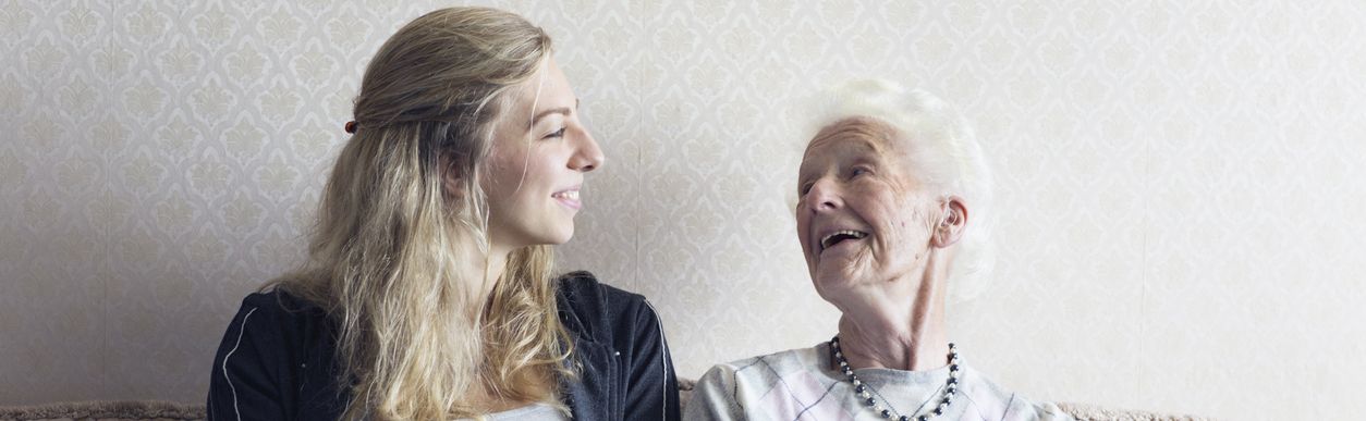 Junge Frau und Seniorin lächeln sich sitzend auf einem Sofa an.