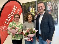 Zwei Frauen und ein Mann stehen nebeneinander vor einer Malteser Fahne. Die Frauen haben einen Blumenstrauß in der Hand.