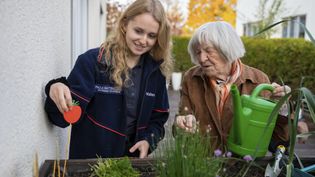 Junge Frau mit dunkelblauer Jacke und ältere Frau mit brauner Jacke gießen und pflegen Pflanzen in einem Hochbeet im Garten.
