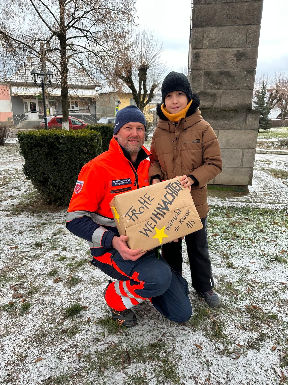 Mann in Malteser Kleidung und Junge in brauner Jacke mit einem Weihnachtspaket auf verschneiter Wiese