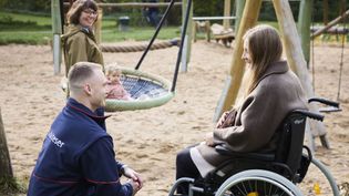 Ein Mann in einer blauen Malteser-Jacke hockt neben einer Frau in einem brauen Mantel, die im Rollstuhl sitzt. Beide sind auf einem Spielplatz. Im Hintergrund schaukelt ein Kleinkind und eine weitere Frau steht neben dem Kind und passt auf.