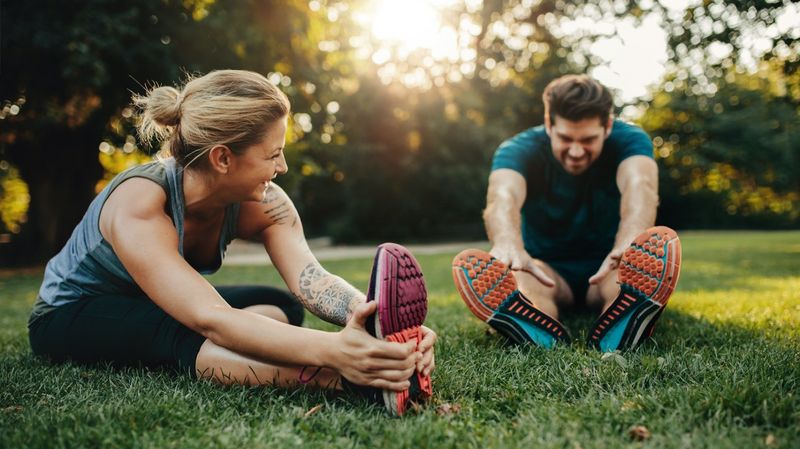 Junger Mann und junge Frau in Sportkleidung machen Dehnübungen auf einer Wiese. 