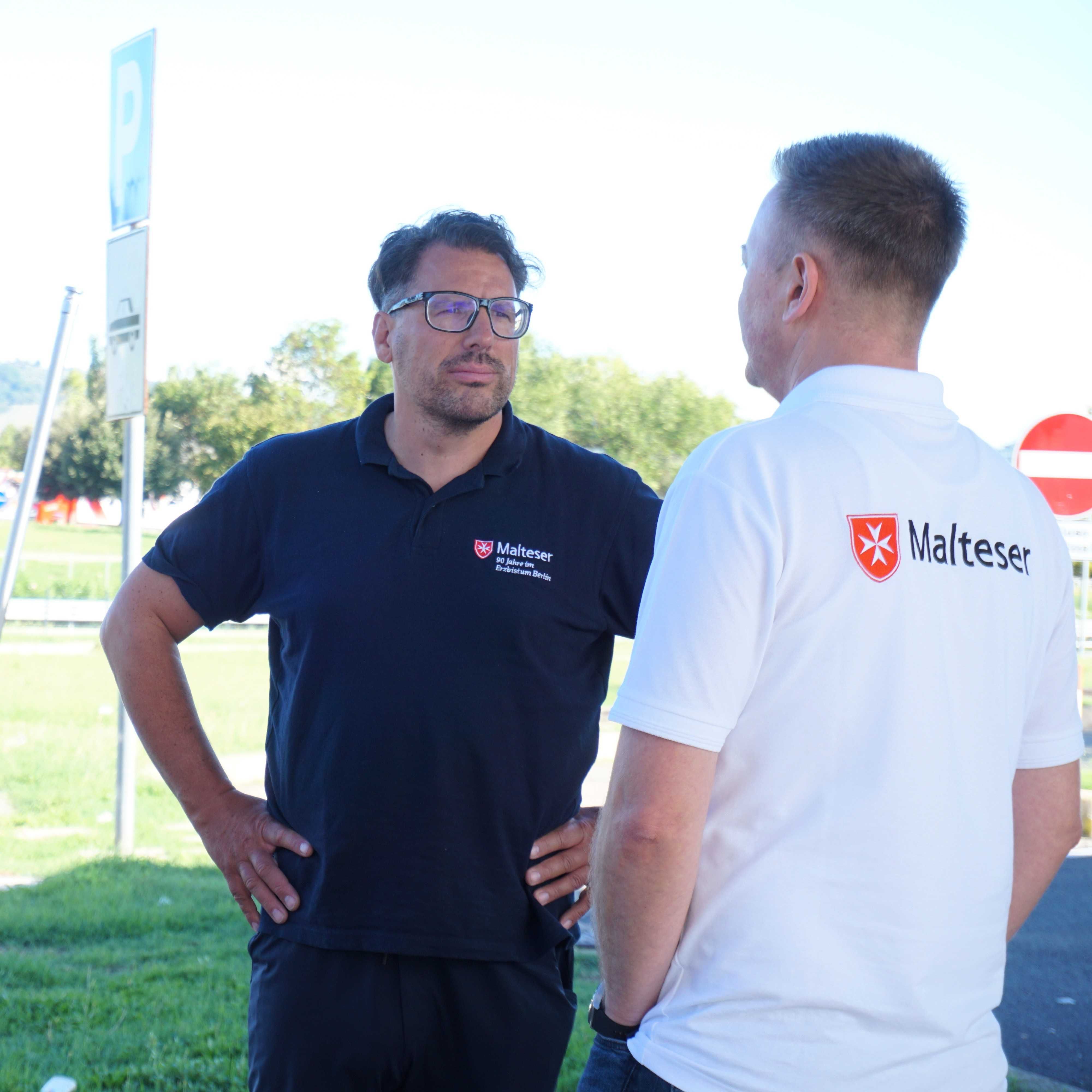 Zwei Männer im Gespräch, einer trägt ein blaues Poloshirt mit Malteser-Logo, der andere ein weißes T-Shirt mit Malteser-Schriftzug und Wappen.