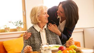 Malteser Mitarbeiterin beugt sich freundlich zu älterer Frau runter, die an einem gedeckten Tisch sitzt