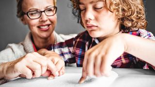Frau sitzt hinter einem Jungen, der in Blindenschrift liest.