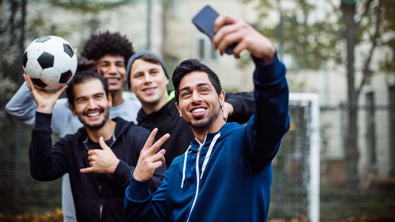 Eine Gruppe junger Männer unterschiedlicher Herkunft steht auf einem Bolzplatz und macht ein gemeinsames Selfie.
