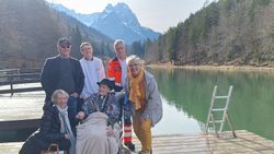 Eine Frau im Rollstuhl umringt von mehreren Personen vor der Kulisse eines Bergsees.