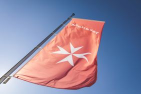 Rote Maltese-Flagge mit weißem Kreuzsymbol an einem Mast vor blauem Himmel.