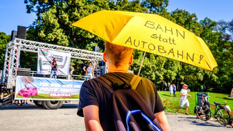 Ein junger Mann auf einer Demonstration hält einen gelben Regenschirm in der Hand, auf dem „Bahn statt Autobahn“ steht. 