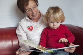 Eine ältere Frau hat ein Kleinkind auf dem Schoß. Beide schauen sich ein Bilderbuch an.