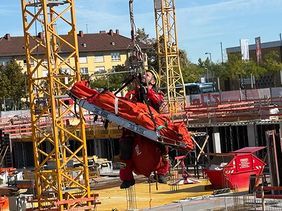 Auf einer Baustelle hängt eine Person in roter Schutzkleidung gesichert an einem Kranhaken. Unter der Person ist eine orangefarbene Trage befestigt. Im Hintergrund sind Kräne, Baugerüste und unfertige Gebäudeteile zu sehen.
